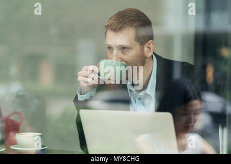 Businessman sitting in cafe, drinking coffee, using laptop, viewed through window Stock Photo