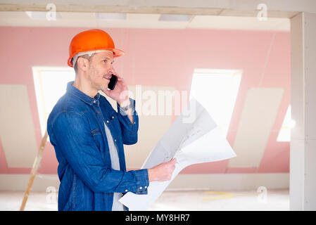 Middle aged construction worker holding plan in his hand while using mobile phone and talking with somebody. Stock Photo