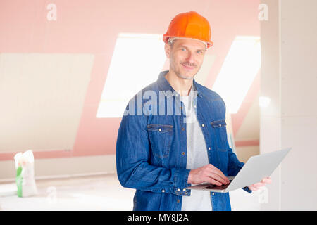 Middle aged construction worker holding laptop in his hand while while standing at constraction site and working. Stock Photo