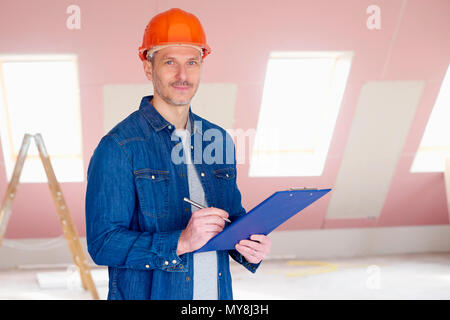 Middle aged handyman wearing hardhat and holding clipboard in his hand while standing at construction site. Stock Photo