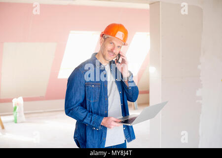 Smiling construction man making call and using his laptop while standing at construction site. Stock Photo