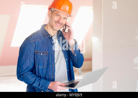 Smiling construction man making call and using his laptop while standing at construction site. Stock Photo