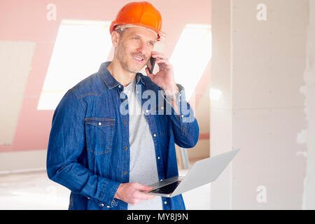 Construction man making call and using his laptop while standing at construction site. Stock Photo