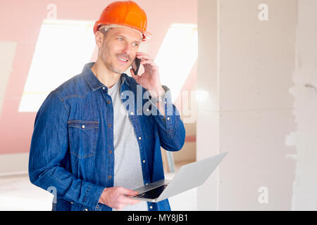 Construction man making call and using his laptop while standing at construction site. Stock Photo