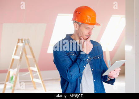 Portrait of middle aged construction worker looking thoughtful while standing at constraction site and using digital tablet. Stock Photo