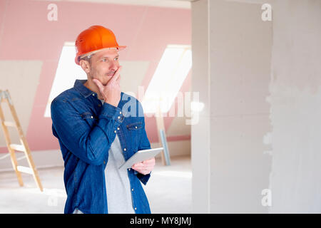 Portrait of middle aged construction worker looking thoughtful while standing at constraction site and using digital tablet. Stock Photo