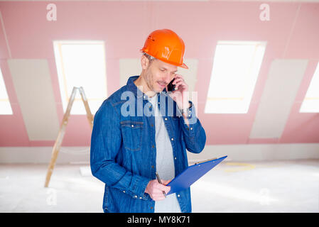 Middle aged handyman wearing hardhat and holding clipboard in his hand while while talking with somebody on mobile phone. Stock Photo