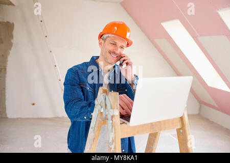 Portrait of happy construction worker using mobile phone and consulting with somebody while using laptop at construction site. Stock Photo