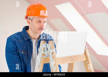 Portrait of middle aged construction worker using laptop while standing on the ladder at construction site. Stock Photo