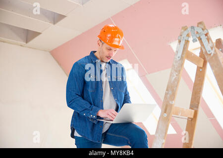 Portrait of middle aged construction worker using laptop while standing on the ladder at construction site. Stock Photo
