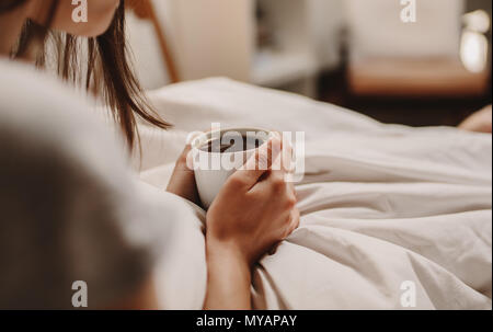 Close up of a woman sitting on bed holding a cup of coffee. Woman with bed coffee in hand sitting on bed. Stock Photo