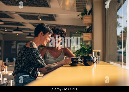 Female friends sitting at coffee shop and smiling. Young woman using mobile phone while sitting with her female friend at coffee shop. Stock Photo