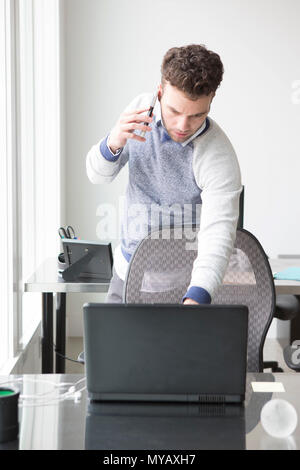 A young businessman checks his laptop while talkingb on a cellphone. Stock Photo