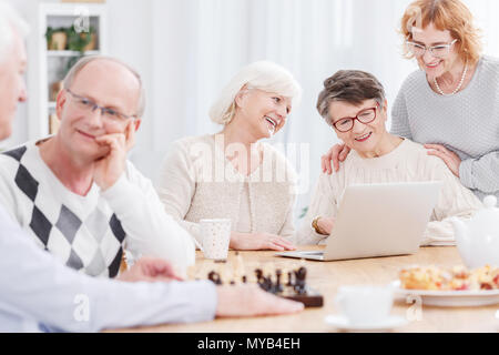 Two older men playing chess and three senior women using laptop Stock Photo
