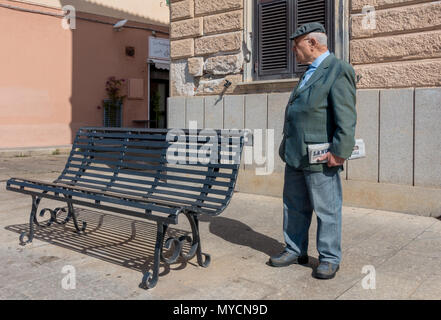 Senior Sardinian man  holding Italian newspaper with hearing aid and flat cap standing by a bench Stock Photo