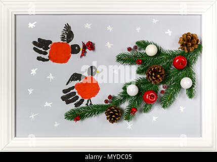 Prints of the hands palms are stylized as bullfinches. Branch of fir tree with cones, red apples and white balls. Handmade picture in a frame as an il Stock Photo