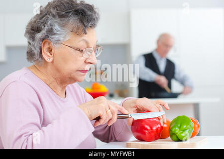 senior woman cutting capsicums while husaband cooks in the background Stock Photo