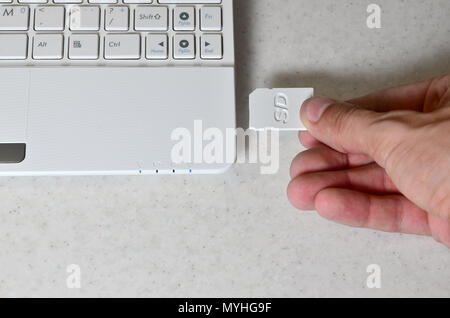 A male hand inserts a white compact SD card into the corresponding input in the side of the white netbook. Man uses modern technologies to store memor Stock Photo