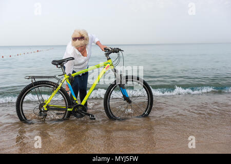 Woman in jeans standing in the water on a beach leaning over a bicycle in the surf in a concept of a healthy outdoor lifestyle Stock Photo