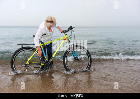 Woman standing in her jeans in the surf leaning over to check her bicycle with gently breaking waves breaking at her feet Stock Photo