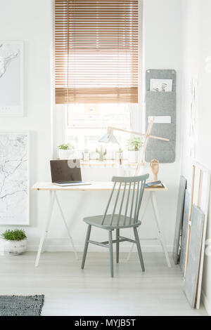 Grey wooden chair standing by the desk with lamp and laptop with empty screen in white room interior with window and fresh plants Stock Photo