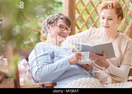 Happy elderly woman drinking tea and nurse reading her a book in the garden Stock Photo