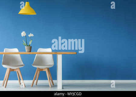 Two chairs, flower, yellow lamp and wooden table on a blue, empty wall in dining room interior Stock Photo