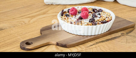 Deep white plate with muesli, dried cranberries, nuts, fresh raspberries and coconut placed on a board on wooden table Stock Photo