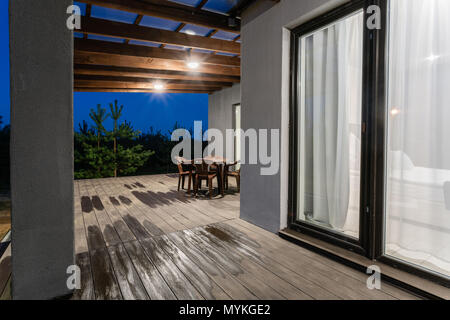 Side view of an open veranda in front of a modern forest cottage. Pine young forest under the sunset rays in the background. Stock Photo