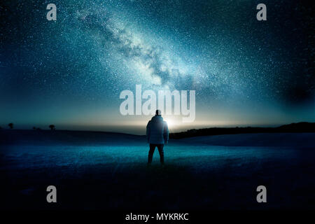 A man stands watching with wonder and amazement as the moon and milky way galaxy fill the night sky. Night time landscape. Photo composite. Stock Photo