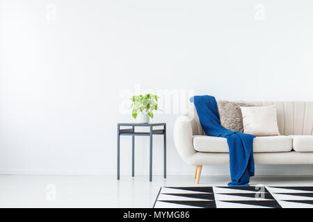 Blue blanket on beige sofa against the wall with copy space in bright living room with plant on cabinet and geometric carpet Stock Photo