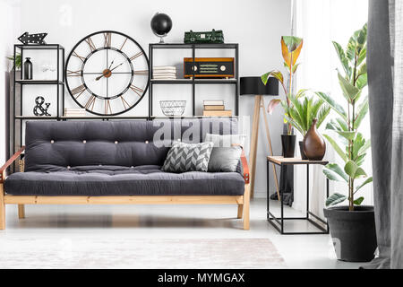 Black wood and leather couch with a patterned cushion standing in bright living room interior with clock on the wall Stock Photo