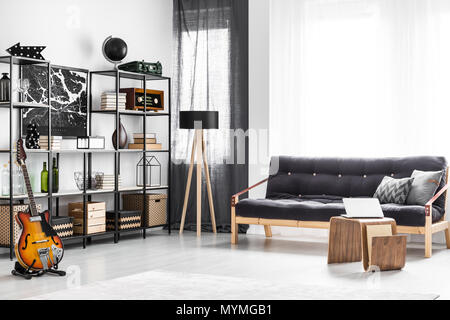 Wooden table and black sofa with cushions in bright guy's room interior with guitar, lamp and shelves Stock Photo