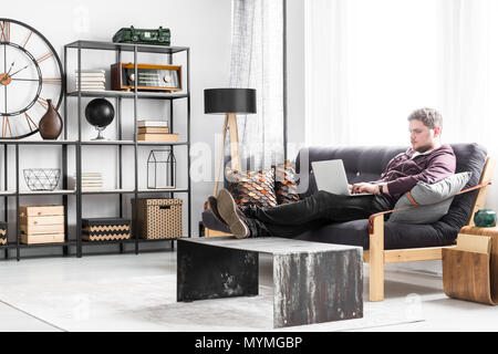 Young man with laptop sitting on the couch in black and white living room interior with industrial table Stock Photo