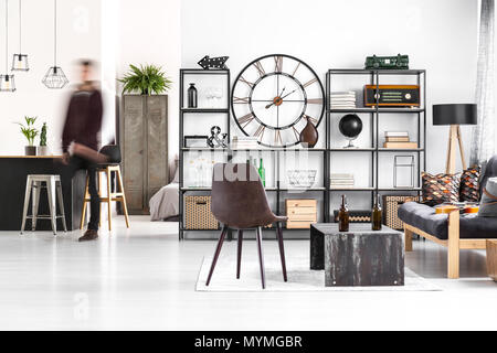 Brown chair and sofa next to a table with bottles in guy's room interior with kitchenette and clock on the wall Stock Photo