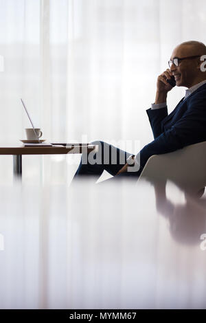 Businessman sitting at office cafeteria and making a phone call. Male executive using mobile phone at office lobby. Stock Photo