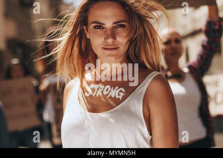 Confident female activist protesting outdoors with group of women in background. Woman with word strong written on her body. Stock Photo