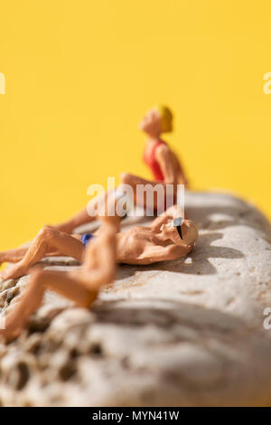 closeup of some different miniature people wearing swimsuit sunbathing on a rock, on a yellow background Stock Photo