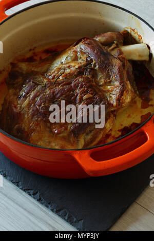 Roasted shoulder of lamb in an orange ceramic roasting dish after cooking on slate and grey wood kitchen worktop Stock Photo