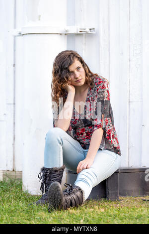 Young woman sitting by side of farm farmhouse building vertical in autumn or fall with boots in park in Virginia thinking in countryside Stock Photo