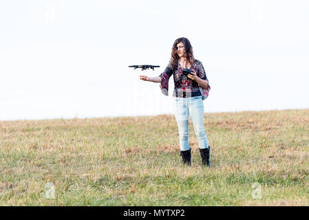 Young woman holding up controlling video camera drone in autumn or fall with boots in park in Virginia countryside open field hill Stock Photo