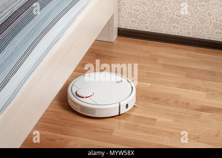 Robot vacuum cleaner runs on wood parquet floor under bed in bedroom. Modern smart cleaning technology housekeeping. Stock Photo
