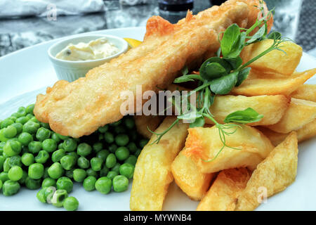 Crispy battered fish and chunky potato chips served with boiled green peas and mayonnaise, traditional English food . Stock Photo