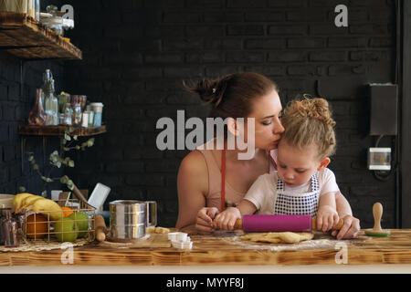 Young mother and daughter prepare cookies in kitchen. They are in aprons. Little girl rolls dough with rolling pin. Mother kisses child, encouraging Stock Photo