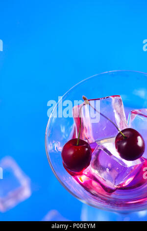 Cherry cocktail close-up. Martini glass with ice cubes and cherries on a bright blue background with copy space. Hot summer day refreshment concept