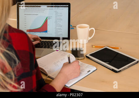 Blonde woman sitting at a laptop and writing. There is a laptop, a tablet, a phone and a notebook on the table. Stock Photo