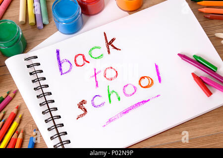 The words Back to School written on a white folded notebook with various paints, crayons and pencils on a school desk. Stock Photo