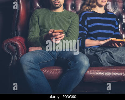 A young couple on a sofa at home are reading and using a mobile phone Stock Photo