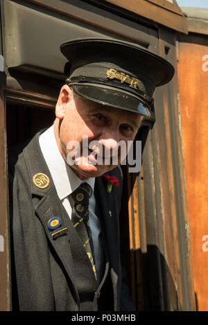 Close-up, front view portrait of senior UK ticket inspector, isolated, in vintage railway carriage, leaning out of train window, greeting passengers. Stock Photo