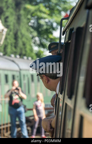 Rear view close up of train conductor (guard) in uniform, on vintage UK steam train, head out of window as train is leaving heritage railway station. Stock Photo